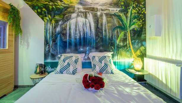 Een slaapkamer met een muurschildering van een waterval, een bed met witte beddengoed, twee gestreepte kussens, een nachtkastje met een lamp en een bos rode rozen.
