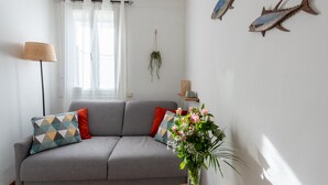 A living room with a grey sofa, patterned cushions, a floor lamp, a vase with flowers, and two wall-mounted fish decorations. - L'Echappée Bleue - Appt pour 3 Vue mer latérale (Ault)