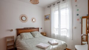 A bedroom with a large bed, a wooden headboard, a bedside table, a small shelf, a mirror, and a window with curtains. - L'Echappée Bleue - Appt pour 3 Vue mer latérale (Ault)