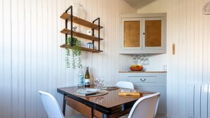 A dining area with a wooden table, white chairs, a shelf with glass jars, and a kitchen area with a cabinet and a fruit bowl. - L'Echappée Bleue - Appt pour 3 Vue mer latérale (Ault)