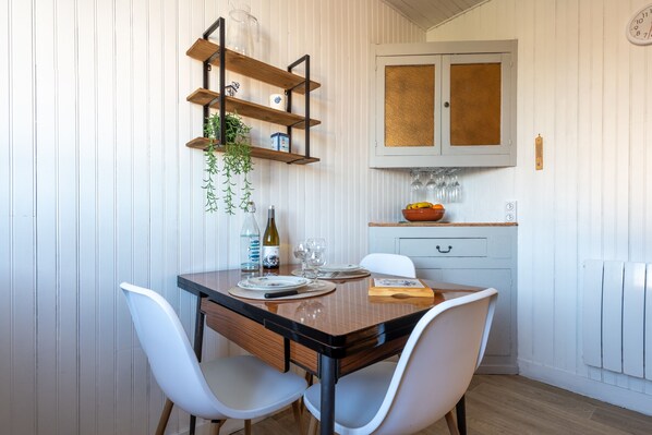A dining area with a wooden table, white chairs, a shelf with glass jars, and a kitchen area with a cabinet and a fruit bowl.
