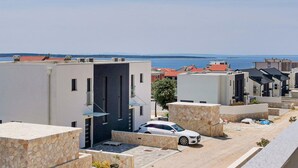 A residential area with modern buildings, a white car parked in front, and a view of the sea in the background.