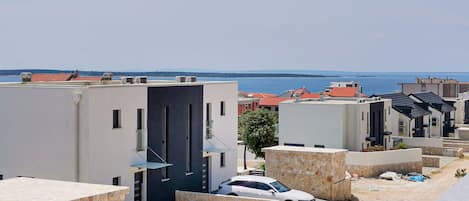 A residential area with modern buildings, a white car parked in front, and a view of the sea in the background.