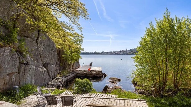 A wooden deck with chairs overlooking a calm lake, a rocky cliff, and a distant town.