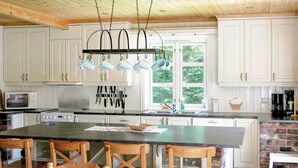 A kitchen with white cabinets, a central island with chairs, a hanging pot rack, and a window with a view of trees.