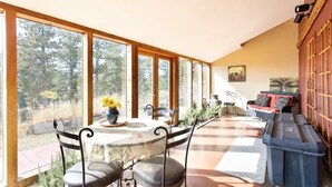 Ein sonnendurchflutetes Zimmer mit Esstisch und Stühlen, einer Couch und Blick auf Bäume durch große Fenster.