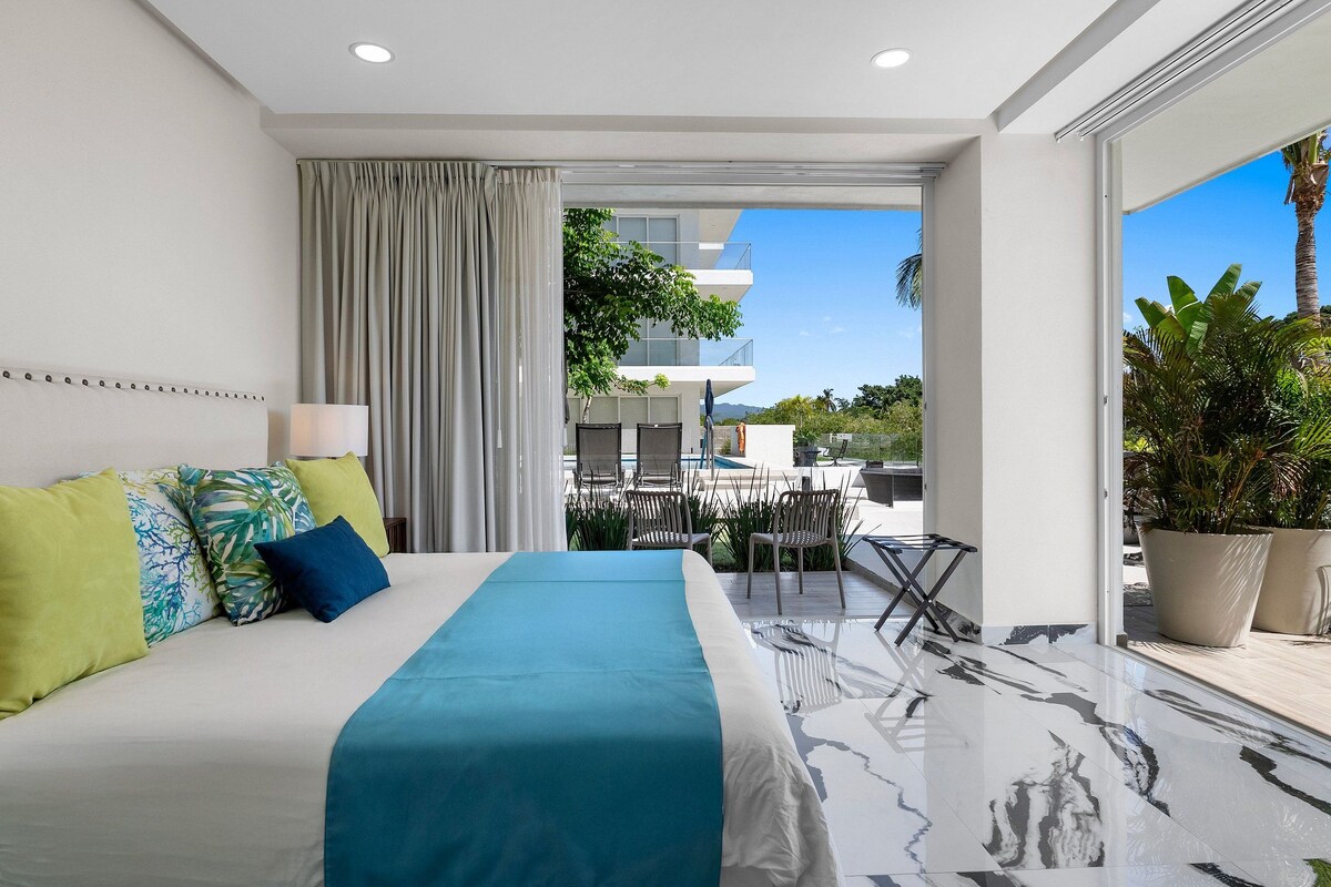 A modern hotel room with a large bed, a balcony with outdoor seating, and a view of the cityscape.