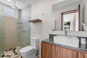 A modern bathroom with a glass shower enclosure, a white toilet, a vanity with a sink, and a mirror. - Quinta San Miguel alberca y playa 5 min (Nuevo Nayarit)