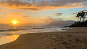 Uma praia com pegadas a conduzir à água, um pôr do sol e palmeiras ao fundo.