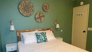 Cama bem arrumada com roupa de cama branca e almofada estampada com folhas, mesa de cabeceira com abajur, e três painéis de parede trançados.