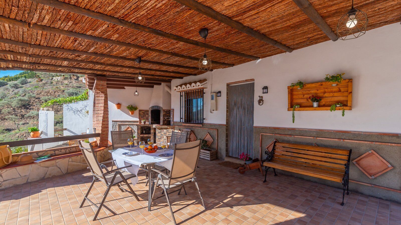 Une terrasse extérieure avec une table ronde, des chaises et un banc en bois. On y trouve également un four en briques et une fenêtre ornée d’une grille décorative.