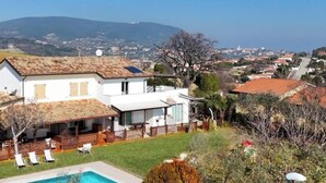 Una casa de dos pisos con piscina, un patio cercado con sillas de descanso y vista a un pueblo y colinas al fondo.