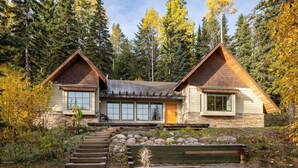 A cabin with a stone foundation, wooden exterior, and large windows. It features a stone staircase leading to the entrance and is surrounded by trees.