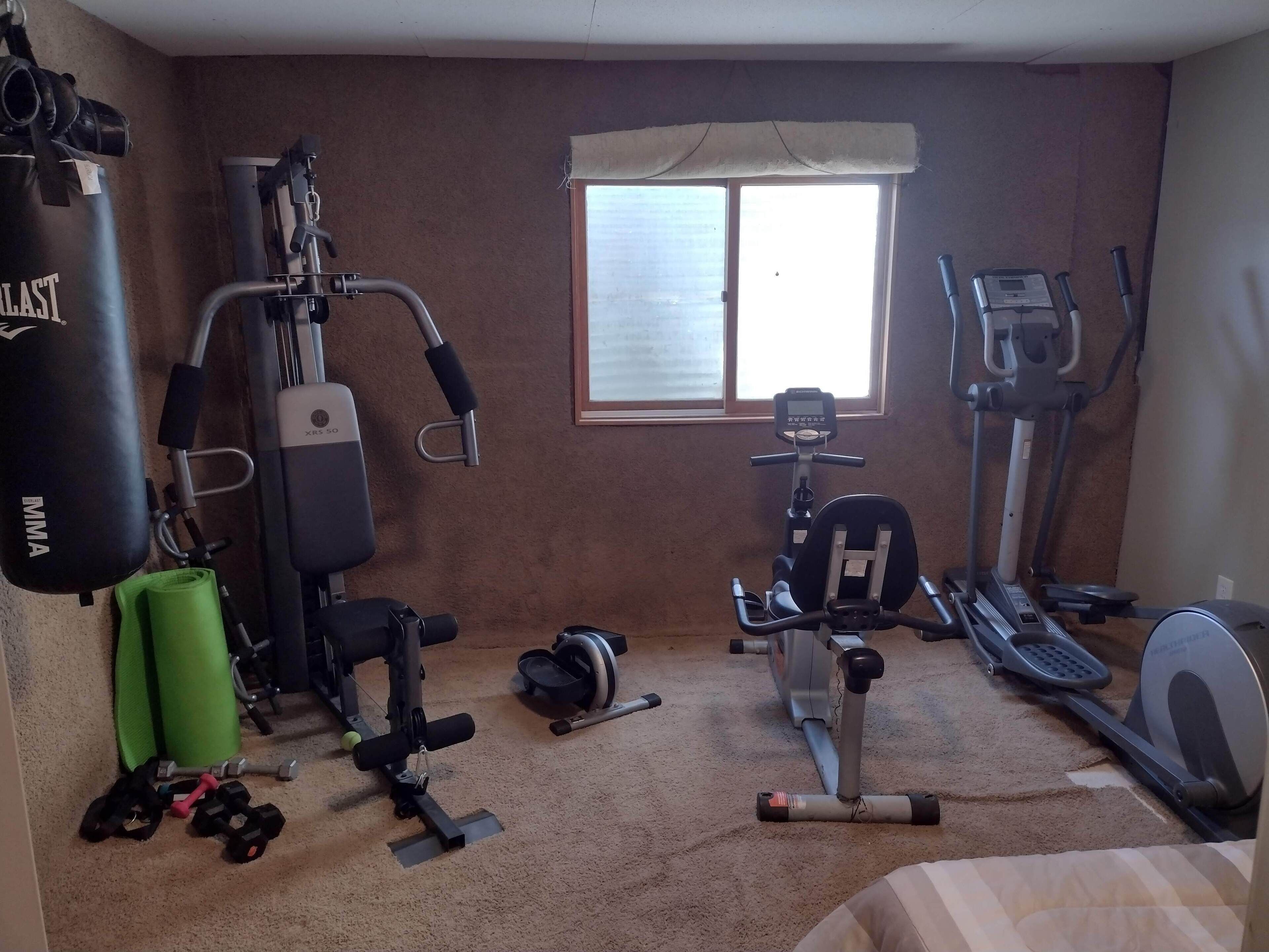 Een huisgym met diverse fitnessapparaten, waaronder een loopband, een hometrainer en krachttoestellen.