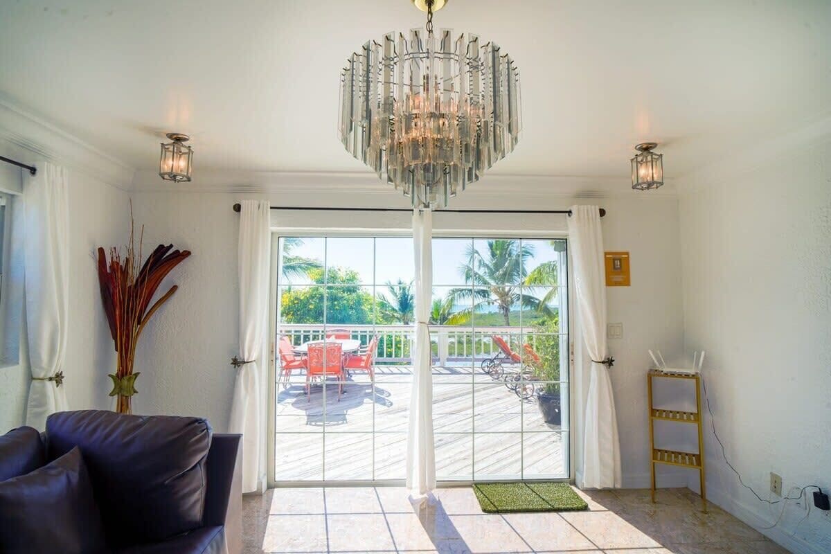 A room with a chandelier, a dining area with red chairs, a view of the outdoors, and a white wall with a power outlet.