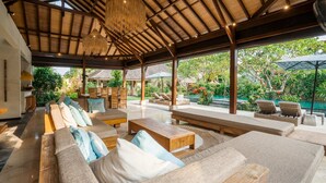 A spacious outdoor lounge area with wooden furniture, a thatched ceiling, and a view of a pool and garden. - Villa Alroda Ayun (Canggu)