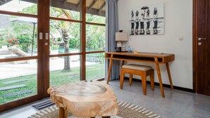 A room with a large sliding glass door leading to a garden, a wooden table with a bench, a desk with a lamp, and a wall-mounted artwork. - Villa Alroda Ayun (Canggu)
