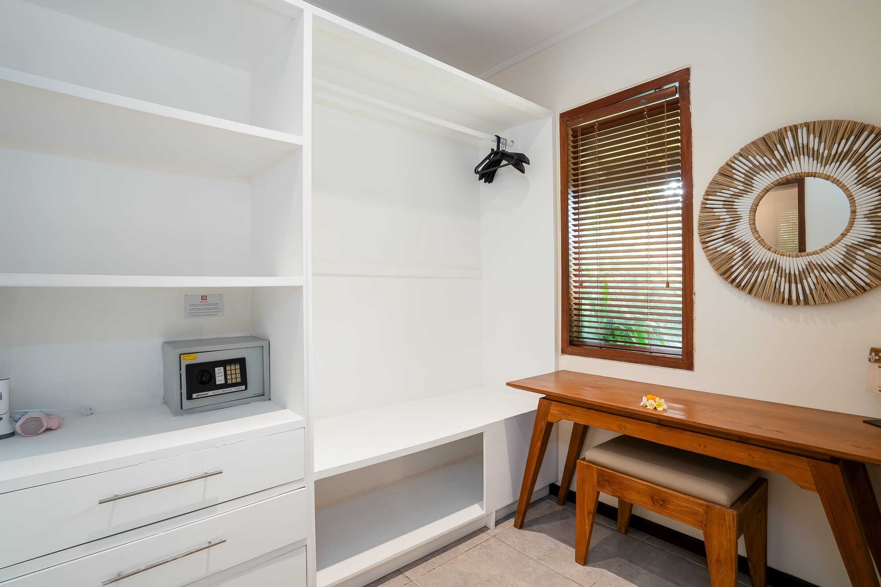 A room with a white wardrobe, a wooden desk, a mirror, and a window with blinds.