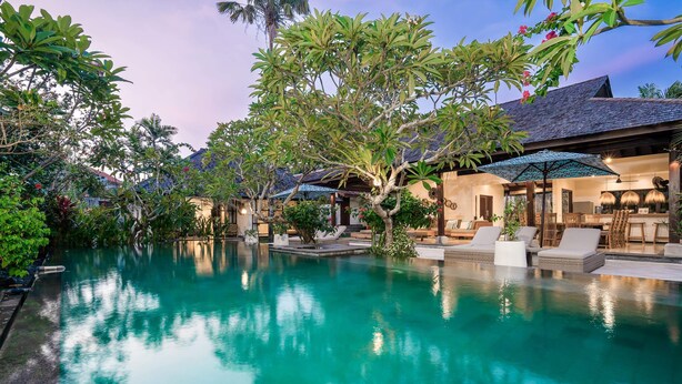 A serene pool area with lounge chairs and umbrellas, surrounded by lush greenery and a traditional building with a tiled roof. - Villa Alroda Ayun (Canggu)