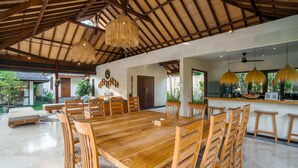 A spacious dining area with a long wooden table and chairs, a thatched ceiling, and a view of the outdoors. - Villa Alroda Ayun (Canggu)