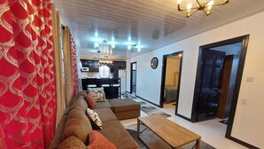 A living room with a sofa, ottoman, and a wooden coffee table. There is a kitchen in the background with a dining area. - Cozy 2-bedroom apartment in central location with WiFi, AC (Demerara-Mahaica)