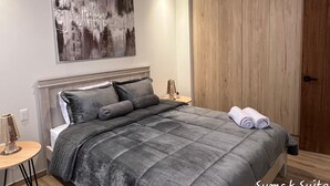 A bedroom with a large bed, a wooden wardrobe, a wall-mounted abstract painting, and a small round table with a lamp.