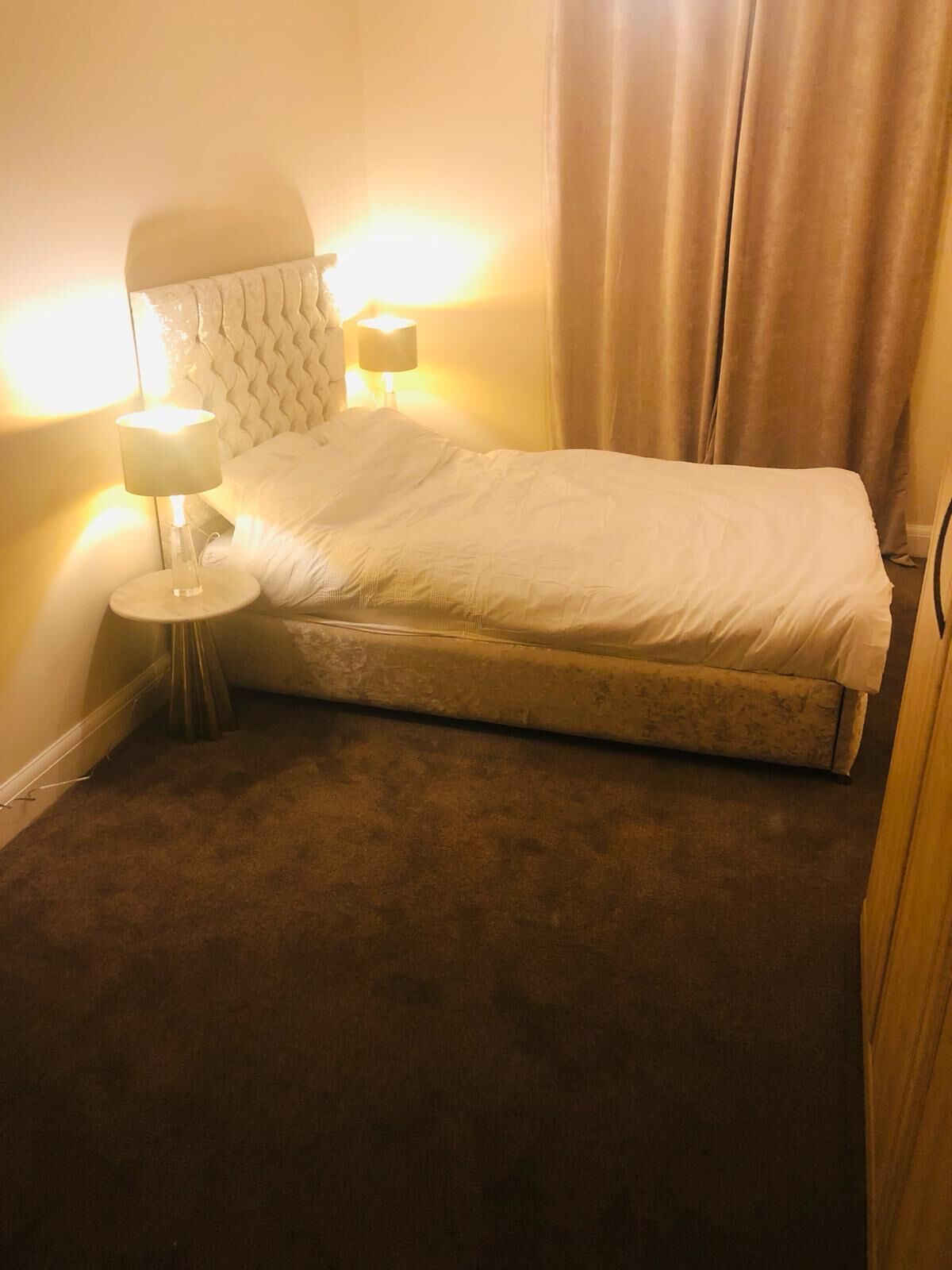A single bed with a white duvet in a room with a brown carpet. There is a bedside table with a lamp and a wall-mounted light.
