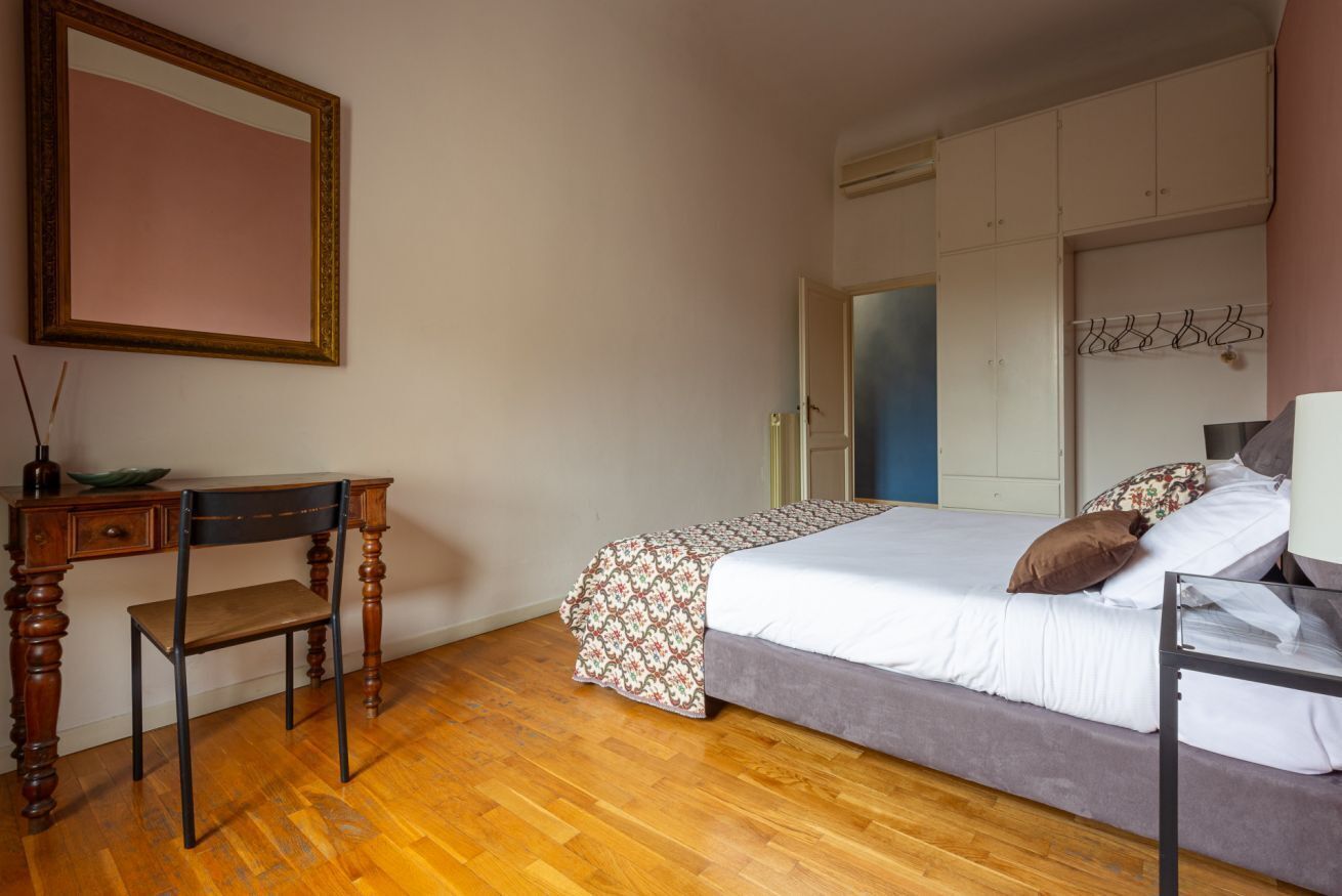 A bedroom with a wooden desk, a chair, a bed with a patterned bedspread, and a wardrobe.