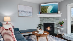 A living room with a fireplace, a television mounted above it, a sofa, a coffee table, and a view of the ocean through the window.