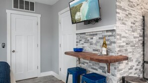 A room with a stone wall, a mounted TV, a wooden bar counter, two blue bar stools, and a bottle of wine. - Studio TC Close to Downtown Beaches and Wineries (Traverse City)