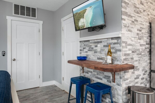 Een kamer met een stenen muur, een wandgemonteerde televisie, een houten bar en twee blauwe barkrukken, plus een fles wijn.