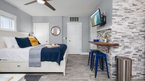 A bedroom with a bed, a ceiling fan, a TV mounted on the wall, and a small kitchenette area with stools and a bar. - Studio TC Close to Downtown Beaches and Wineries (Traverse City)