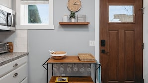 A kitchen with a wooden shelf holding a clock, a bowl, and a basket. There is a door with a window and a wall-mounted light switch. - Studio TC Close to Downtown Beaches and Wineries (Traverse City)