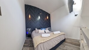 A modern bedroom with a herringbone-patterned headboard, a bed with a beige blanket and pillows, a wooden floor, and a TV mounted on the wall.