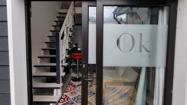 A modern entryway with a glass door labeled "Ok Loft." Inside, there is a staircase leading to a loft-style living area with a patterned rug and a red chair.