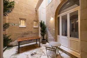 A stone courtyard with a bench, a potted plant, a table with chairs, and a door with glass panels. - Harbour Mansion 2 bedroom Maisonette (Birgu)