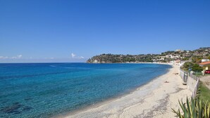 Ein Küstenstrand mit klarem, blauem Wasser, einem sandigen Ufer und einer Hügelsiedlung mit Gebäuden.