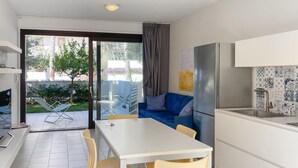 Eine moderne Küche mit weißer Arbeitsplatte, einem eingebauten Backofen und einem Edelstahlkühlschrank. Es gibt eine Essecke mit weißem Tisch und Stühlen, und eine blaue Couch im Hintergrund.