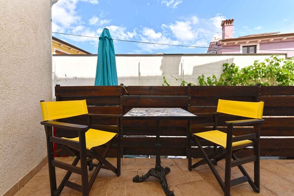 A patio with a wooden table and two chairs, a blue umbrella, and a view of a building with a pink facade.