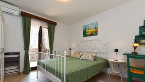 Un dormitorio con cama blanca, ropa de cama verde, un cuadro en la pared, una mesita de noche con una lámpara y un radiador.