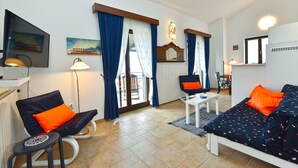 A modern living room with a sofa, a chair, a small table, and a TV. There is a window with curtains, a mirror, and a radiator. - Holiday apartment with pool, barbecue and fitness equipment for shared use (Barbariga)