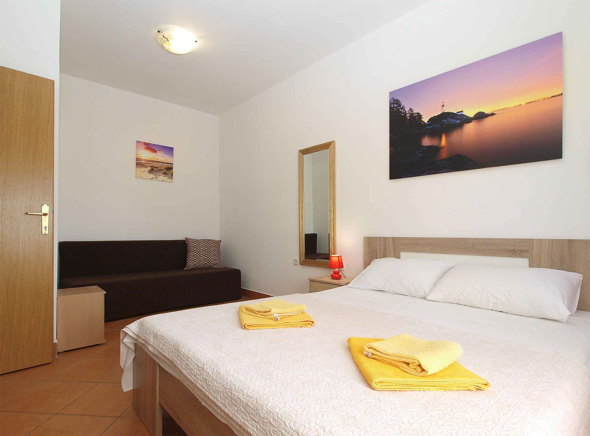 Ein Hotelzimmer mit einem Bett, einem Nachttisch mit Lampe, einem Spiegel und einem Sonnenuntergangsbild an der Wand.