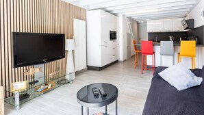 A modern living room with a flat-screen TV, a glass coffee table with magazines, a sofa, and a kitchen area with a dining table and chairs. - Maison mitoyenne proche du lac ! (BISCARROSSE)