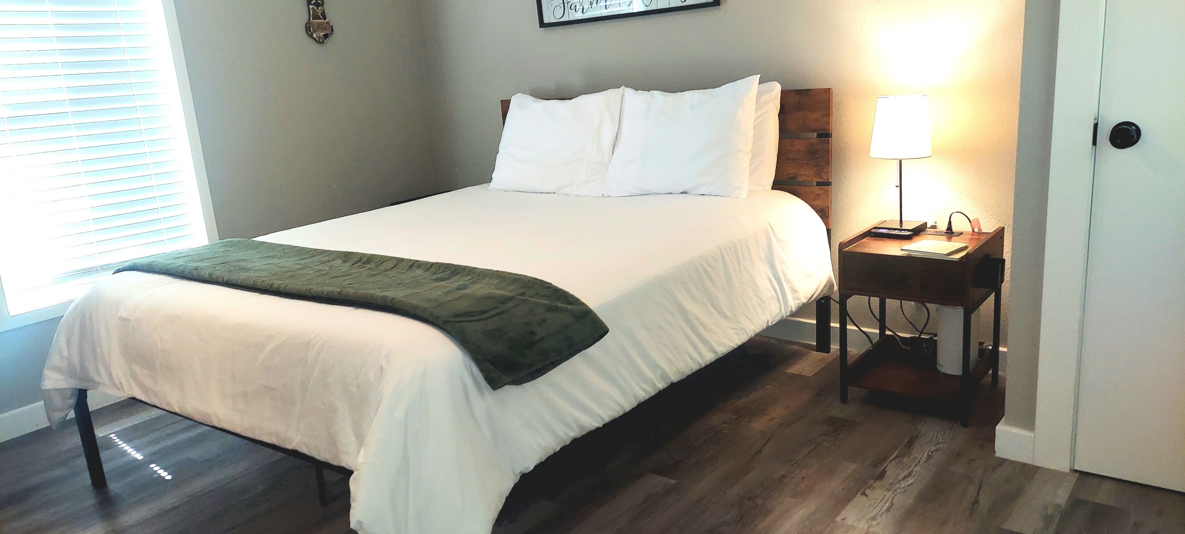 Un lit bien fait, avec du linge de lit blanc et un plaid vert, une table de chevet avec une lampe et une tête de lit en bois.