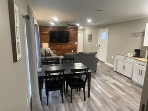 A modern kitchen with white cabinets, a dining table with chairs, a gray sofa, and a TV mounted on the wall.