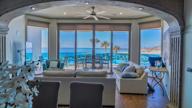 Amplia sala de estar con un gran ventanal con vista a una piscina y playa, un ventilador de techo y un diseño interior moderno.