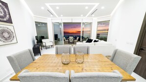 Un comedor moderno con una mesa de madera y sillas grises tapizadas, vistas de la ciudad a través de puertas de vidrio, y un sofá blanco al fondo.