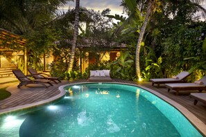 A swimming pool with wooden deck and lounge chairs, surrounded by lush greenery and a well-lit building in the background. - Casa das Águas Trancoso (Porto Seguro)