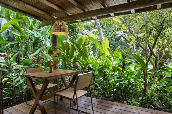 A wooden deck with a table and chairs, surrounded by lush tropical plants and a woven hanging light fixture. - Casa das Águas Trancoso (Porto Seguro)