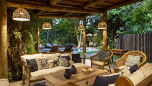 Um pátio externo com móveis de vime, uma mesa de madeira e luminárias de vime pendentes.